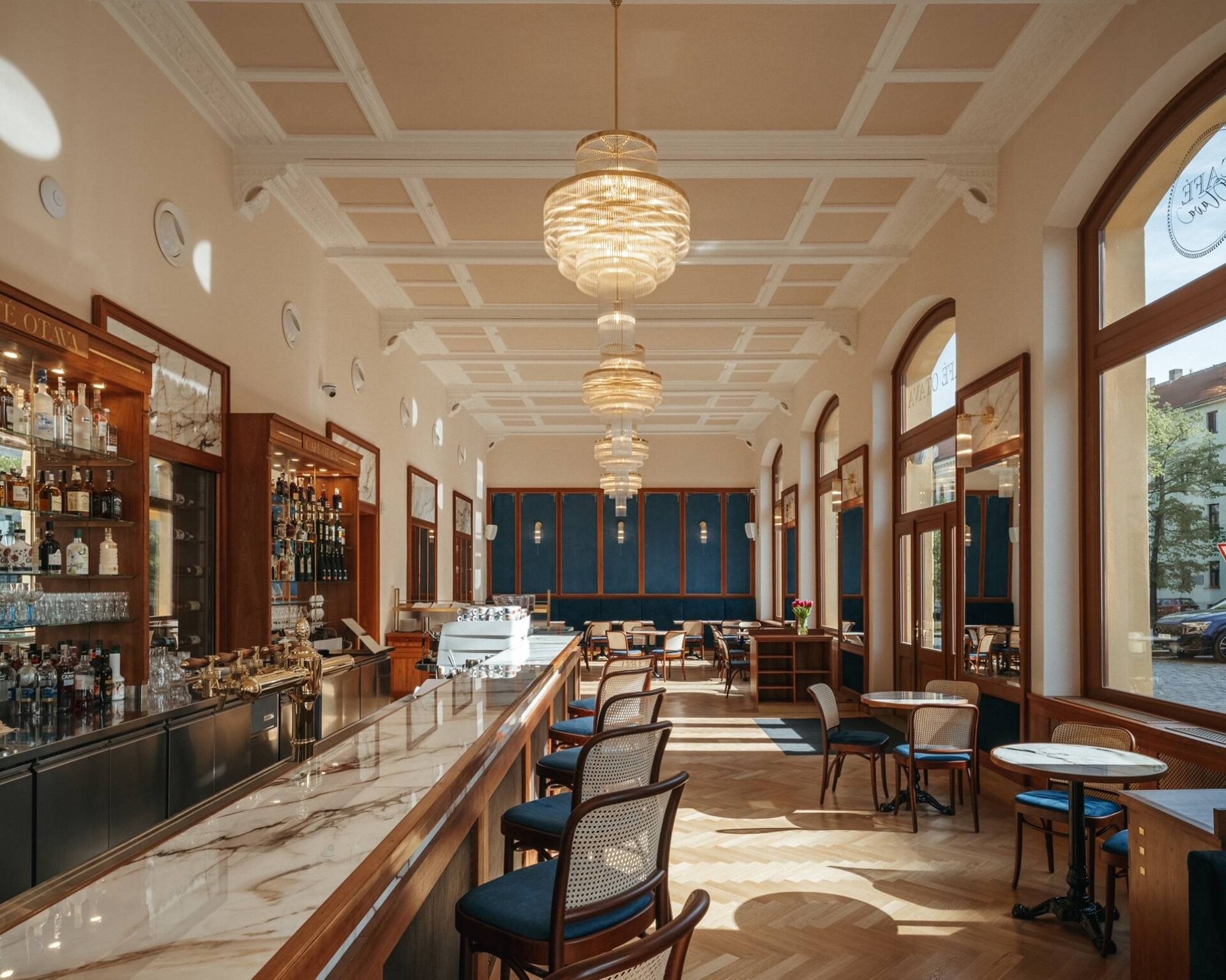 Vstupný bar Café Otava s mramorovou doskou, dreveným barovým pultom a tromi mosadznými lustrami pod kazetovým stropom